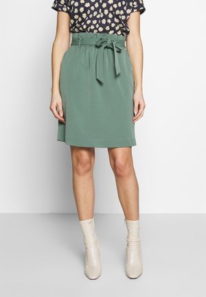 Woman wearing a knee-length green skirt with a tied belt, floral blouse, and off-white ankle boots, standing against a plain white background.