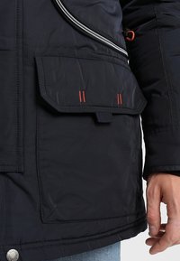 Gros plan d'une main reposant près d'une poche de veste marine avec un rabat, des coutures orange et une fermeture éclair visible au-dessus de la poche.