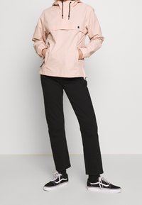 Jasnoróżowy polarowy płaszcz z kapturem, pół-zamek z przodu i kieszeń kangurza. W zestawie z czarnymi spodniami i czarnymi butami Vans.