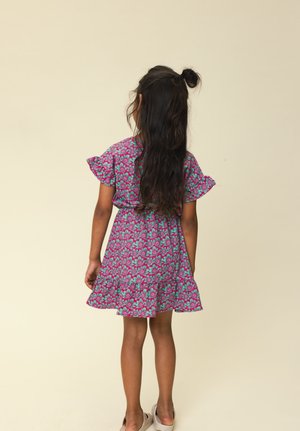 Jong meisje met lang donker haar in een bloemenjurk van roze en groen, staand met haar rug naar de camera tegen een effen beige achtergrond.