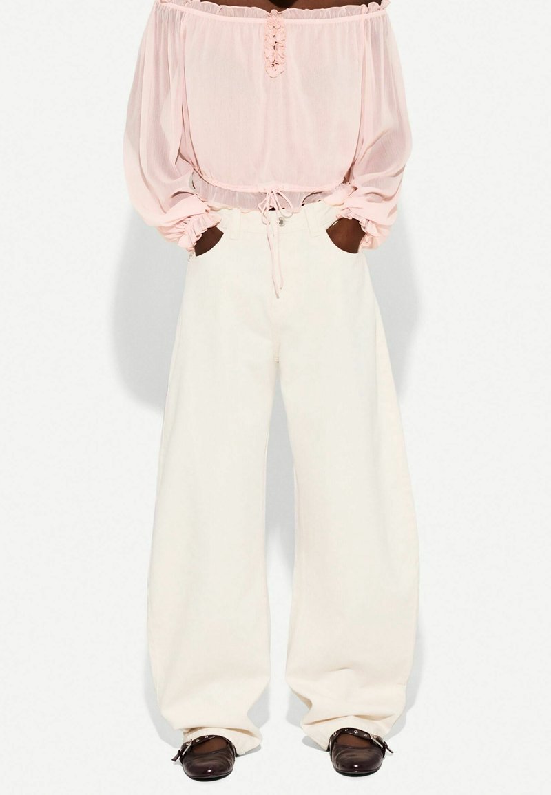 Personne portant un chemisier rose clair épaules dénudées à manches longues, un pantalon blanc large et taille haute, et des chaussures marron foncé avec des boucles.