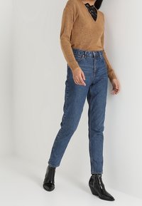 Maglione marrone a maglia con scollo a V e dettagli in pizzo, indossato sopra jeans blu a vita alta. Stivali neri con zeppa completano l'outfit.