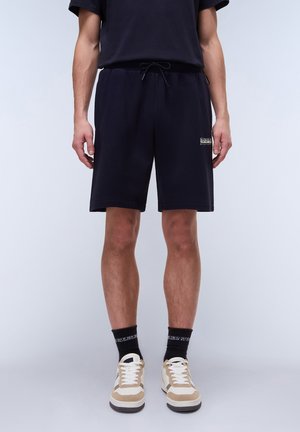 Navy cotton shorts with an elastic waistband, drawstring, side pockets, and small logo detail on the left leg. Paired with beige sneakers.