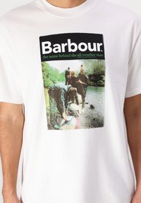 Tricou din bumbac alb, cu un print grafic ce reprezintă oameni lângă apă, cu text negru și verde care citește "Barbour" și "numele din spatele bărbatului pregătit pentru orice vreme."