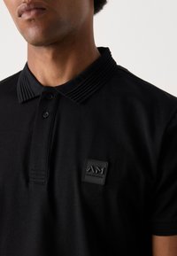 Zwarte poloshirt met een gestructureerde kraag, drie knopen en een opvallende logopatch op de borst. Gladde stof met een aansluitende pasvorm.