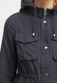 Navy blue hooded jacket with zip closure, two front pockets, drawstring waist, and gold-tone hardware details. Smooth fabric texture.