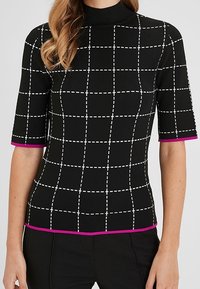 Černý pletený top s krátkým rukávem a bílým mřížkovým vzorem, rolákem a růžovým lemem na rukávech a okraji, nošený s černými kalhotami.