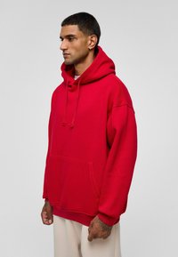 Rode sweatshirt met capuchon, voorzak en geribbelde manchetten. Glad materiaal, oversized ontwerp. Model staat tegen een neutrale achtergrond.