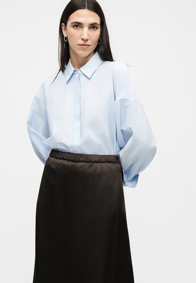 Γυναίκα με μακριά σκούρα μαλλιά, που φοράει κρίκους στα αυτιά, ένα φαρδύ ανοιχτό μπλε πουκάμισο με κουμπιά και μια μαύρη φούστα με ψηλή μέση, που στέκεται σε λευκό φόντο.