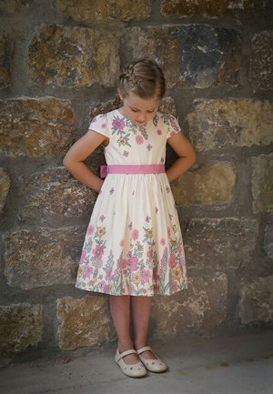 Junges Mädchen mit geflochtenem Haar, das ein weißes Blumenkleid mit rosa Gürtel und weißen Schuhen trägt, steht vor einer Steinmauer und schaut nach unten.