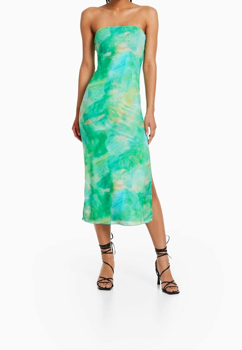 Femme portant une robe midi sans bretelles, en tie-dye vert et bleu avec une fente sur le côté, associée à des sandales à talons hauts noires à lanières.
