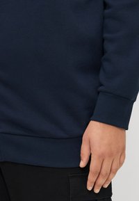 Navy blue sweatshirt with ribbed cuffs and hem, featuring a soft texture. Includes a hand resting on the side, against a black background.