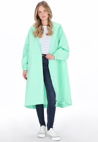 Mentaszínű kapucnis esőkabát laza szabással, enyhén hosszabb alsó szegéllyel és elasztikus mandzsettával, csíkos felső és kék farmer fölött viselve.