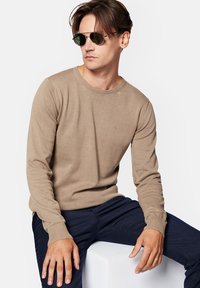 Beżowy sweter z dzianiny z okrągłym dekoltem, długimi rękawami i ściągaczami, w połączeniu z ciemnogranatowymi spodniami; model nosi zielone okulary przeciwsłoneczne.