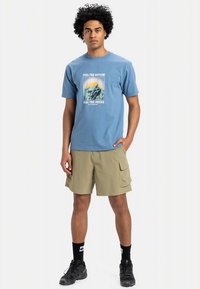 Zils kokvilnas T-krekls ar kalna grafiku un tekstu. Bēša krāsas kargo šorti ar kabatām. Melnas zeķes un kurpes noslēdz apģērbu.