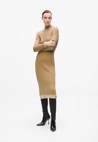 Abito in maglia color beige con colletto alto, texture a costine e logo in evidenza sull'orlo. Abbinato a pantaloni neri aderenti e stivali neri lucidi.