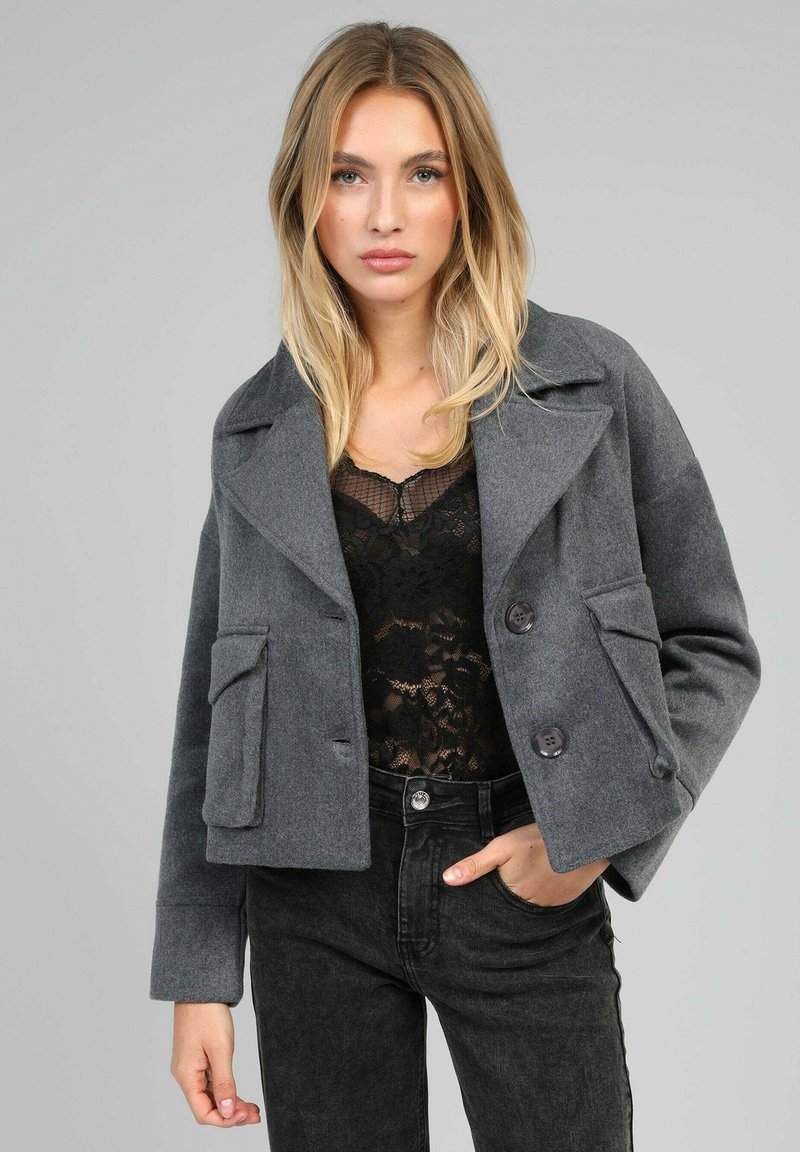 Veste en mélange de laine grise avec un col cranté, deux poches avant et fermeture à double bouton. Portée par-dessus un haut en dentelle noir et un jean noir.