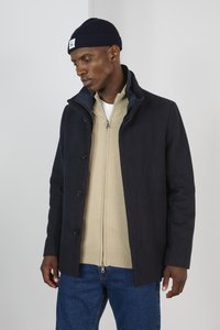 Cappotto in misto lana blu navy con colletto alto e cinque bottoni. Indossato sopra una giacca leggera beige con zip e jeans blu. Accompagnato da un berretto blu navy.