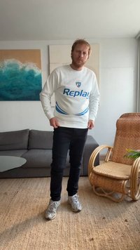 Valge särk, millel on sinine "Replay" logo; mustad teksad ribakanga tekstuuriga; hallid tossud. Seisab heleda tausta ees.