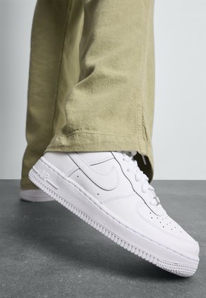 Białe sneakersy Nike Air Force 1 noszone z beżowymi spodniami, stopa lekko uniesiona nad szarym teksturowanym tłem podłogi.