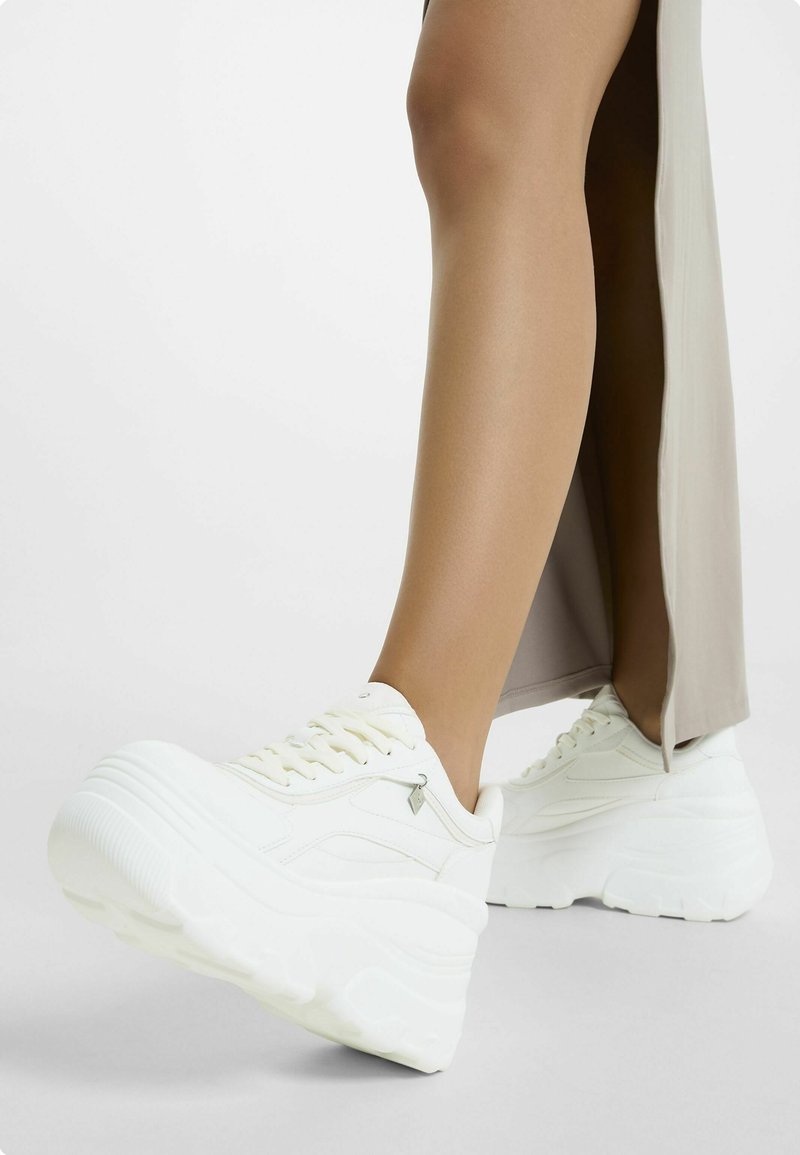 Sneakers platform bianchi con suola texturizzata, design con lace-up e un accento del logo discreto sul lato, abbinati a un indumento beige chiaro.