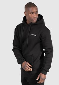 Homme en hoodie noir avec poche avant et logo "Smilodox" blanc regarde à sa gauche devant un fond gris uni.