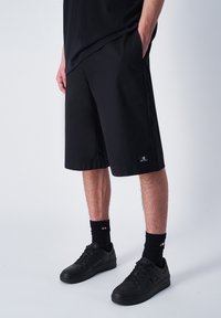 Zwarte shorts met een losse pasvorm en boven de knie lengte, gemaakt van lichtgewicht stof. Voorzien van een logopatch en zijzakken. Zwarte sneakers en sokken.