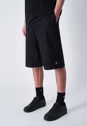 Shorts noirs à coupe ample, arrivant au-dessus du genou, fabriqués en tissu léger. Équipés d'un patch logo et de poches latérales. Baskets noires et chaussettes.