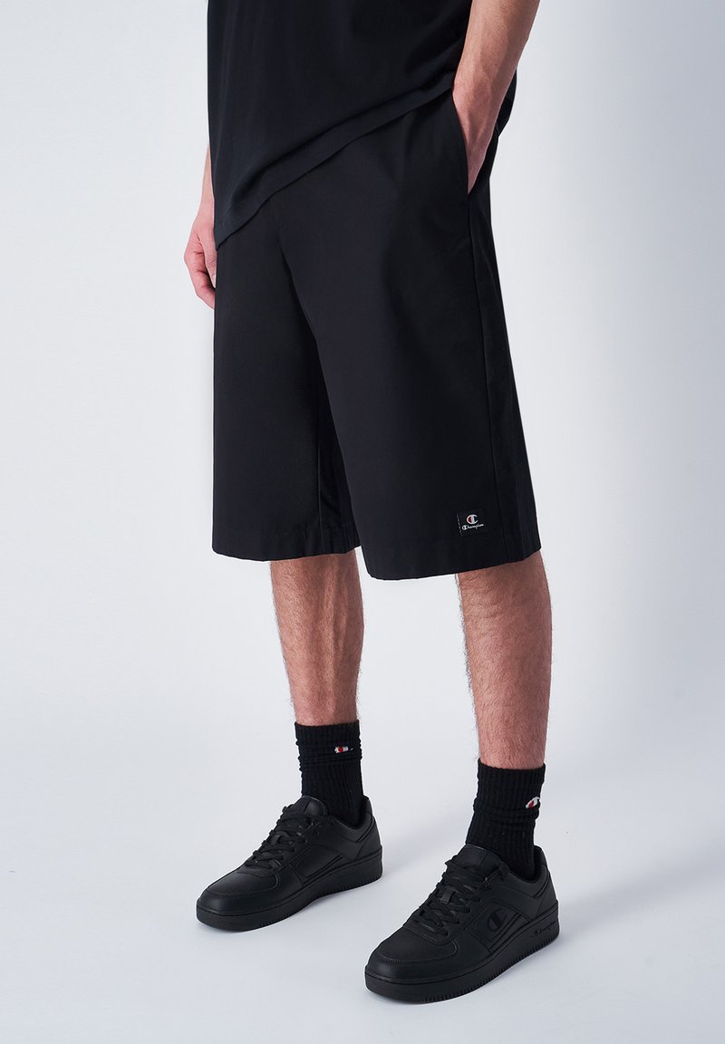 Zwarte shorts met een losse pasvorm en boven de knie lengte, gemaakt van lichtgewicht stof. Voorzien van een logopatch en zijzakken. Zwarte sneakers en sokken.