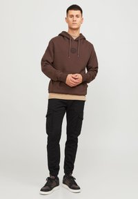 Brun huvtröja med dragsnören och framficka, lager på en tan-färgad skjorta. Svarta cargobyxor med sidofickor och åtsittande manschetter. Sneakers med bruna detaljer.