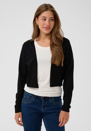 Jonge vrouw met lang bruin haar die een zwarte korte cardigan draagt over een wit shirt en blauwe spijkerbroek, kijkt iets naar de zijkant en glimlacht.