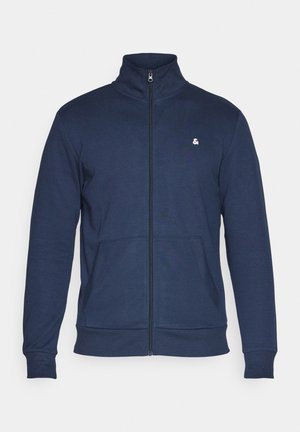 Tumesinine tõmblukuga jakk kõrge kaelusega, kahe esimest taskuga, ribidega mansetid ja alläär, valmistatud pehmest kangast, millel on rindkerel väike valge logo.