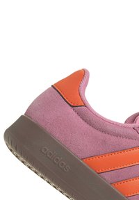 Rosa mockaläder Adidas-sneakers med orange accenter och brun sula, som visar texturerat mockaläder och präglad logotyp på sulan.