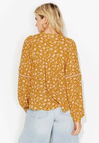 Femme aux cheveux blonds portant des boucles d'oreilles en or et un blouse à manches longues à fleurs jaune moutarde, associé à un jean bleu clair, vue de dos.