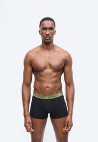 Atlétikus férfi áll előre nézve, fekete boxeralsót visel, olívazöld derékpánttal, amelyen a "BOSS" felirat látható, egyszerű szürke háttér előtt.