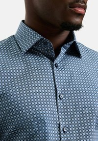 Buttons-up Hemd aus blauer Stoff mit geometrischem Muster in verschiedenen Schattierungen. Kurzer Kragen und zwei Knöpfe an der Vorderseite sichtbar.