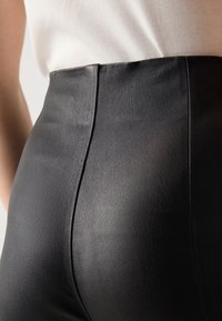 Detailní záběr osoby, která má na sobě černé kožené kalhoty s vysokým pasem a bílé žebrované tričko zastrčené dovnitř.