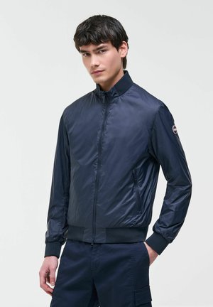 Joven con una chaqueta bomber azul marino con cremallera, puños y cuello acanalados, posando con una mano en el bolsillo contra un fondo liso.