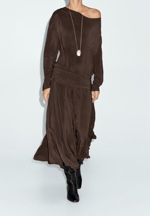 Femme portant une longue robe marron épaules dénudées avec une taille froncée et des bottes noires montant jusqu'aux genoux, accessoirisée d'un long collier pendentif blanc.