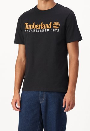 Mann trägt ein schwarzes T-Shirt mit kurzen Ärmeln, auf der Brust ist das in Braun und Weiß gestickte Timberland-Logo mit der Aufschrift „Timberland Established 1973“.