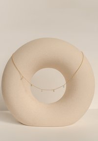 Collana in oro con più ciondoli, esposta su un supporto circolare beige texturizzato. I ciondoli includono piccoli elementi decorativi.