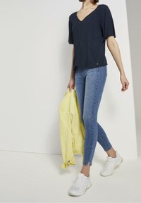 Donna che indossa un top a V blu navy, jeans skinny azzurri con fessure alla caviglia, scarpe da ginnastica bianche, con una giacca gialla nella mano sinistra.