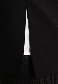 Un tissu noir au toucher lisse présente une fente sur le côté, révélant une doublure blanche en dessous. Un tissu noir plissé est visible en dessous.