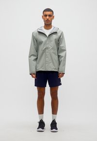 Άνδρας που στέκεται, φοράει ανοιχτό γκρι μπουφάν με κουκούλα, άσπρο πουκάμισο, σκούρα μπλε σορτς, άσπρες κάλτσες και μαύρα αθλητικά παπούτσια σε λευκό φόντο.