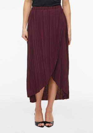 Femme portant une jupe portefeuille plissée bordeaux avec des talons ouverts noirs, debout devant un fond blanc uni.