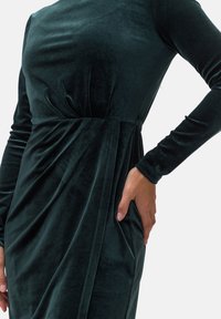 Czarna zielona sukienka z aksamitnego materiału o dopasowanej górnej części, długich rękawach i drapowanym efekcie w talii. Gładka tekstura i minimalistyczny design.