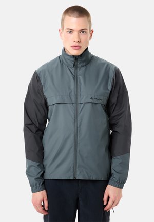 Giovane uomo che indossa una giacca Vaude grigia e nera con cerniera e polsini elasticizzati, in piedi davanti a uno sfondo chiaro e uniforme.