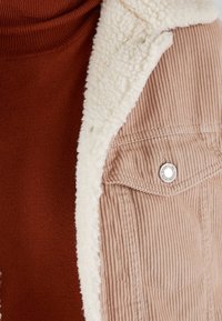 Giacca in corduroy marrone con fodera in sherpa panna. Presenta un bottone argentato su una tasca sul petto. Sotto, un dolcevita color ruggine.