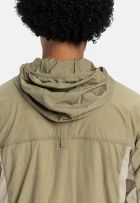 Beige letvægtsjakke med hætte, der har tekstureret stof og et diskret logo. Inkluderer rygventilation og stingdetaljer.