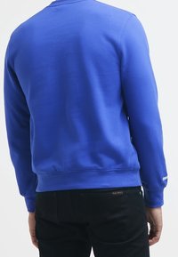 Uomo che indossa una felpa a maniche lunghe blu semplice e pantaloni neri, visto da dietro, in piedi su uno sfondo bianco.